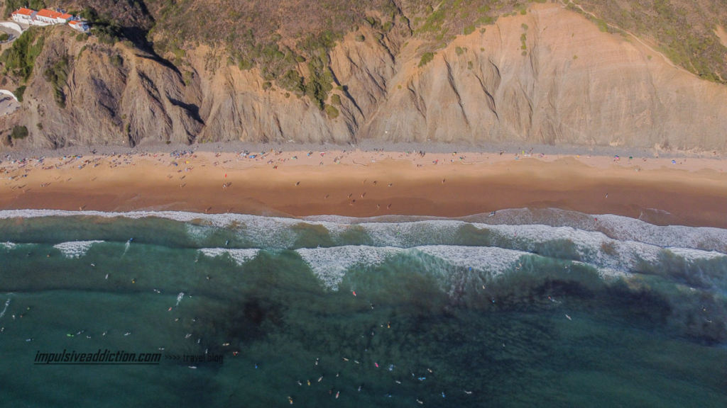 Um mar cheio de surfistas, na praia da arrifana: pequenos pontos coloridos junto à praia.