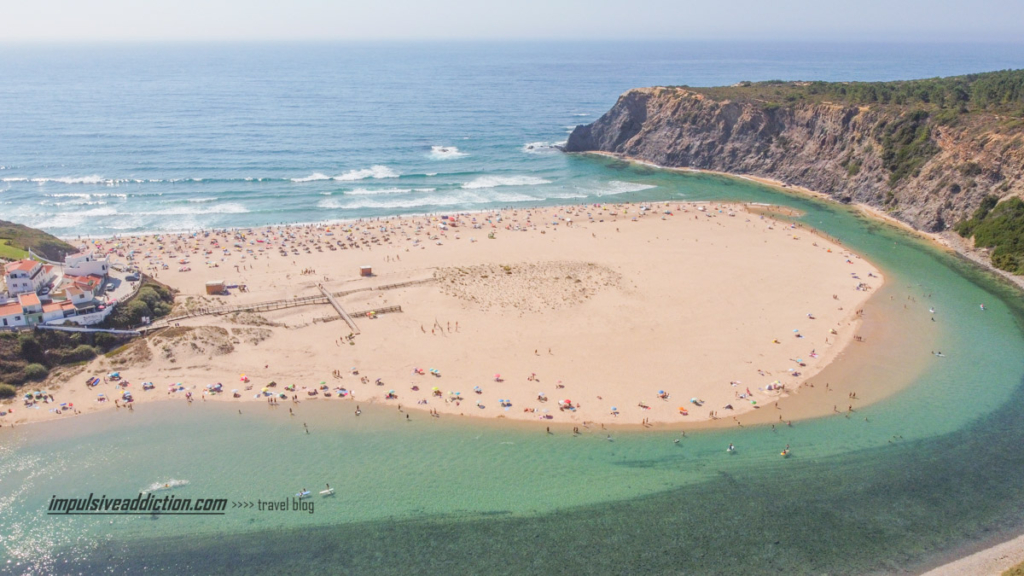 Praia de Odeceixe, vista da lateral, com a ribeira de Seixe a contorná-la em direção ao Mar. As pessoas ocupam o areal em redor da lingua da areia junto ao mar ou rio.