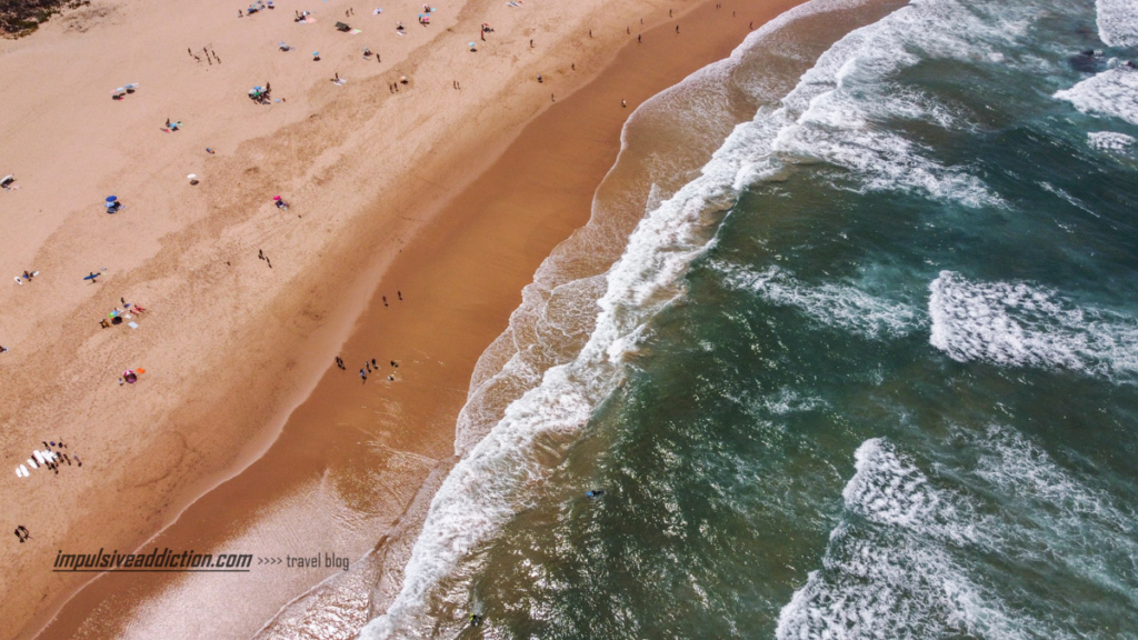 Praia de Monte Clérigo com areal em tons laranja e água do mar em tons de verde escuro. Pessoas espalhadas pelo areal.